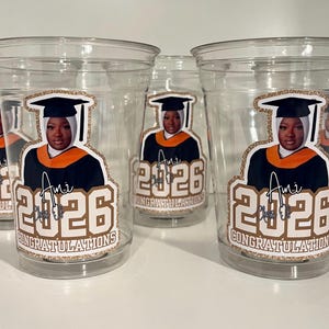 May include: Clear plastic cups featuring a sticker of a graduate wearing a black cap and gown with an orange stole. The sticker includes the text 'Ami', 'Class of 2026', and 'CONGRATULATIONS' in a gold glitter-style border on the front of each cup.