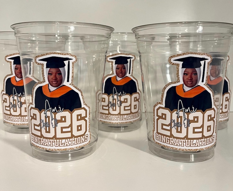 May include: Clear plastic cups featuring a sticker of a graduate wearing a black cap and gown with an orange stole. The sticker includes the text 'Ami', 'Class of 2026', and 'CONGRATULATIONS' in a gold glitter-style border on the front of each cup.