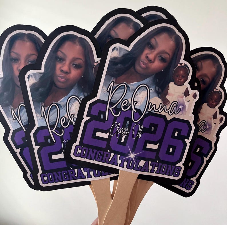 May include: A stack of graduation photo props featuring a young woman. The props are black and purple with the text "Rekonna Class Of 2026 Congratulations". Each prop has a wooden handle.