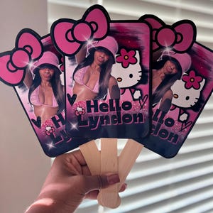 May include: A set of three pink and black Hello Kitty-themed hand fans. Each fan features a photo of a person wearing a pink bikini and hat, with the text "Hello Lyndon". The fans have a pink bow at the top and wooden handles.