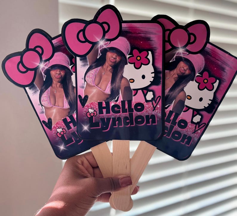 May include: A set of three pink and black Hello Kitty-themed hand fans. Each fan features a photo of a person wearing a pink bikini and hat, with the text "Hello Lyndon". The fans have a pink bow at the top and wooden handles.