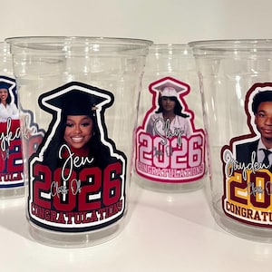 May include: Four clear plastic disposable cups featuring personalized graduation decals. Each cup displays a portrait of a graduate in a cap and gown, the name of the graduate, the year 2026, and the word CONGRATULATIONS in bold lettering.