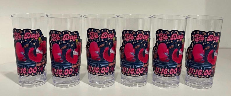 Pu&ograve; includere: Sei bicchieri in plastica trasparente disposti in fila, ognuno con una decalcomania grafica con il testo "Ye Day" e "11:16:00" in rosa, insieme a un'illustrazione stilizzata di una persona in abito rosso seduta su una superficie scura con bordo leopardato.