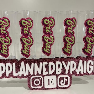 Pu&ograve; includere: Cinque bicchieri in plastica trasparente, ognuno con una decalcomania verticale con il testo Bri Day in lettere verde lime su sfondo magenta glitterato. Un cartello glitterato magenta mostra il testo bianco PPLANNEDBYPAIGE con icone.