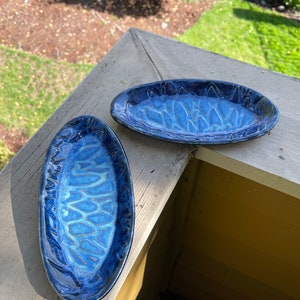 May include: Two handmade, oval-shaped ceramic dishes with a glossy, deep blue glaze. The dishes have a textured, organic pattern on the interior, with variations in the blue tones. The dishes are displayed outdoors on a wooden surface.