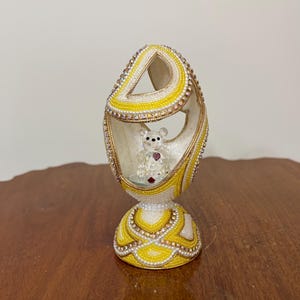 以下が含まれることがあります： 装飾的な卵型の装飾品。卵は主に白、黄色、金で、複雑なビーズ細工が施されており、内部には小さな透明なテディベアのフィギュアが入っています。ベースは黄色と白で、詳細なパターンがあります。