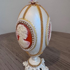 以下が含まれることがあります： カメオの肖像画が施された、白い卵型の装飾品。金色のトリム、パール、クリスタルのトッパーで飾られています。ベースは白で、葉のようなアクセントとラインストーンが付いています。ユニークな装飾品。