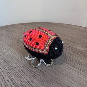 Puede incluir: Figura de mariquita con cuentas, con cabeza negra y cuerpo rojo y naranja. El cuerpo tiene lunares negros y está decorado con cuentas doradas y moradas. La figura se apoya sobre una base transparente con forma de flor.