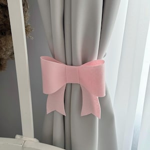 Könnte beinhalten: Ein hellrosa Schleifen-Vorhanghalter aus Filz. Die Schleife befindet sich auf einem grauen Vorhang und sorgt für eine dekorative Note. Ein schlichtes, elegantes Wohnaccessoire.