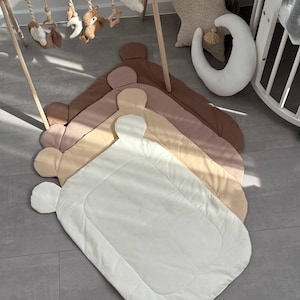 Puede incluir: Una pila de alfombras de juego para bebés en tonos marrones y beige, con una alfombra blanca encima. Las alfombras tienen un diseño único en forma de oso. En el fondo hay un gimnasio de madera para bebés con juguetes colgantes.