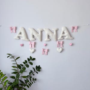 May include: A white felt banner spelling "ANNA" with pink bows and hearts. The letters are puffy and attached to a string, with decorative bows and hearts in pink and white. A green plant is in the lower left corner.