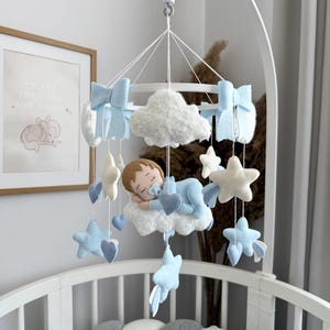Può includere: Un mobile per bambini bianco con una nuvola blu e bianca, un bambino che dorme, stelle e cuori. Il mobile è appeso a una struttura metallica bianca. Il mobile è etichettato "Baby Joys".