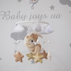 May include: A white and beige baby mobile featuring a teddy bear sleeping on a crescent moon, white clouds, and gold glitter stars. The mobile is attached to a white circular base with a music box that reads "Baby joys" on it.