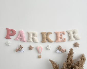 May include: A felt banner spelling "PARKER" in various pastel colors, with star and teddy bear accents. The banner includes a hot air balloon and is designed for nursery or children's room decor.