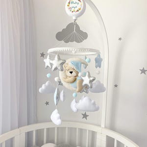 May include: A baby mobile with a sleeping bear on a crescent moon, surrounded by stars, clouds, and pom-poms. The mobile is suspended above a crib. The text "Baby Joys" is on the top of the mobile.
