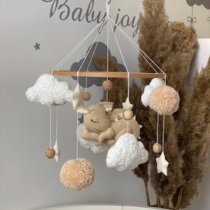 Könnte beinhalten: Ein Baby-Mobile mit einer braunen Holzbasis und weißen Wolken, einem beigen Pompon, einem schlafenden braunen Drachen und einem beigen Stern. Das Mobile hängt an einer weißen Decke.