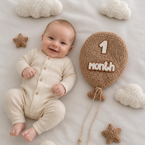 Könnte beinhalten: Ein lächelndes Baby in einem cremefarbenen Strampler liegt auf einer weißen Oberfläche, umgeben von Plüschwolken und sternförmigen Dekorationen. Eine braune Ballon-Requisite zeigt den Text "1 month".