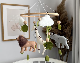 May include: A baby mobile featuring felt animal figures: a lion, giraffe, and elephant, along with white clouds and green leaves. The mobile hangs from a wooden ring, suspended above a crib. The color palette includes beige, brown, white, and gray.