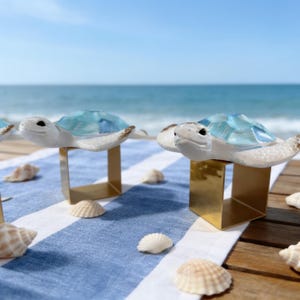 May include: Two napkin rings featuring white sea turtle figures with translucent light blue shells. Each turtle is mounted on a square, polished gold-tone metal base. The design highlights the contrast between the white textured turtle bodies and the smooth, glossy blue shell accents.