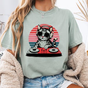 May include: A light green t-shirt with a graphic of a cat wearing headphones and sunglasses while mixing music on a DJ mixer. The cat is in front of a sunset background.