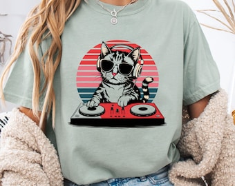 Op de afbeelding: Een lichtgroen T-shirt met een afbeelding van een kat met een koptelefoon en zonnebril, die muziek mixt op een DJ-mixer. De kat staat voor een zonsondergangachtergrond.