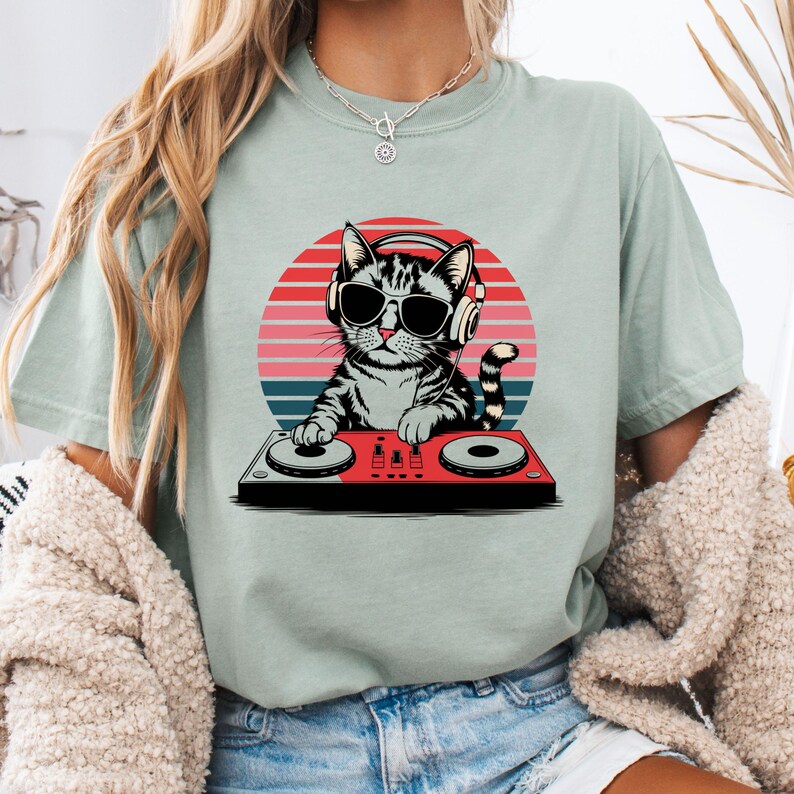 May include: A light green t-shirt with a graphic of a cat wearing headphones and sunglasses while mixing music on a DJ mixer. The cat is in front of a sunset background.