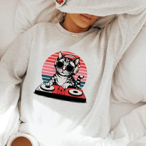 May include: A gray sweatshirt with a graphic of a cat wearing sunglasses and headphones, mixing music on a DJ mixer. The cat is in front of a red, orange, and blue sunset.