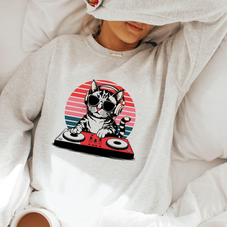 May include: A gray sweatshirt with a graphic of a cat wearing sunglasses and headphones, mixing music on a DJ mixer. The cat is in front of a red, orange, and blue sunset.