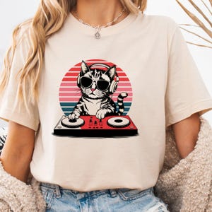 May include: A beige t-shirt with a graphic of a cat wearing headphones and sunglasses while mixing music on a DJ mixer. The cat is in front of a red, orange, and blue sunset.
