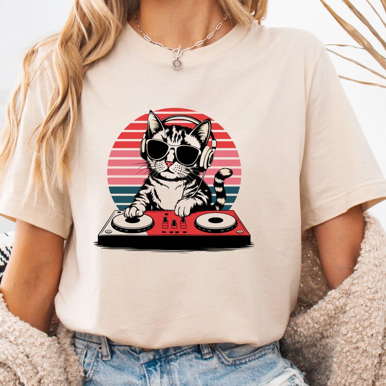 May include: A beige t-shirt with a graphic of a cat wearing headphones and sunglasses while mixing music on a DJ mixer. The cat is in front of a red, orange, and blue sunset.