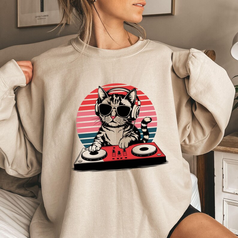 May include: A beige sweatshirt with a graphic of a cat wearing headphones and sunglasses, holding a DJ mixer in front of a sunset background.