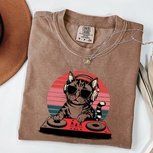 May include: A brown t-shirt with a vintage-style graphic of a cat wearing sunglasses and headphones while DJing. The cat is in front of a red, orange, and yellow sunset.