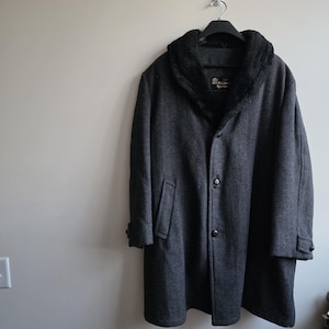 May include: A charcoal grey herringbone coat featuring a black faux fur shawl collar, two front buttons, and two side pockets. The garment includes a black interior lining and a label near the collar that reads The Quilted Sportswear.