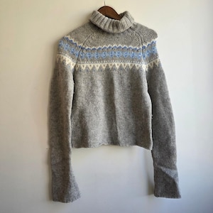 Op de afbeelding: Een grijze coltrui met lange mouwen en een fair isle-patroon op de borst en schouders in lichtblauwe en crèmekleurige tinten. Het gebreide kledingstuk heeft een geribbelde kraag en boorden en hangt aan een houten kledinghanger.