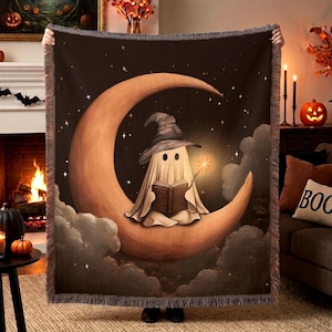May include: A woven throw blanket featuring a dark night sky background with a large orange crescent moon. A white ghost wearing a pointed hat sits on the moon, reading an open book while holding a glowing wand. The edges have a fringed border.