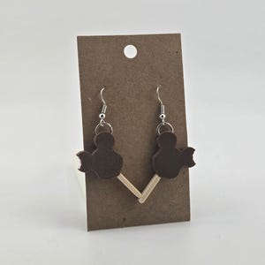 May include: A pair of brown Mickey Mouse-shaped earrings with silver hooks. The earrings are attached to a brown cardboard display card. The earrings are designed to look like chocolate ice cream bars on a stick.