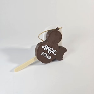 May include: A brown Mickey Mouse-shaped ornament with the word "MAGIC" and the year "2026" in white script. The ornament has a bite taken out of it and is attached to a white popsicle-shaped object with a gold string for hanging.