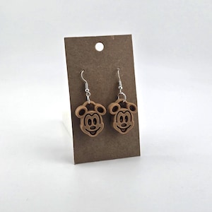 May include: A pair of wooden Mickey Mouse earrings with silver-colored hooks. The earrings are light brown and feature a smiling Mickey Mouse face with black details. The earrings are displayed on a brown card.