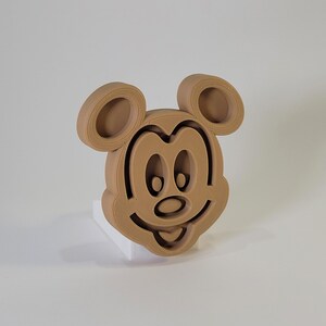 May include: A brown 3D printed figurine of Mickey Mouse's head. The figurine is on a white base.