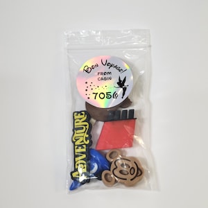May include: A clear plastic bag filled with various Disney-themed charms. The charms include a red ship, a blue hat, a brown Mickey Mouse head, and a yellow charm with the word "Adventure" on it. A holographic sticker with the text "Bon Voyage! From Cabin 7056" and a Tinkerbell image is also included.