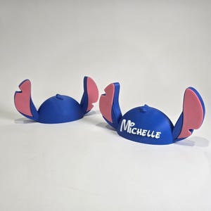 May include: Two blue character-themed headbands with pink accents. One headband has the name "Michelle" in white. The headbands are designed to resemble a popular animated character. The headbands are likely made of plastic.