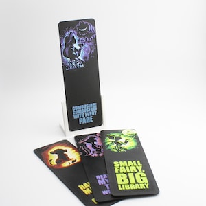 May include: Black bookmarks with colourful illustrations and text. One bookmark features Alice in Wonderland with the text "Curiouser and Curiouser with every page." Other bookmarks have fairy and silhouette designs with text.