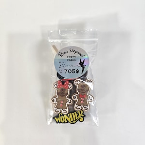 May include: A clear plastic bag containing two gingerbread cookie figures, one with a red bow and the other with a red bow tie. The bag has a circular sticker with the words "Bon Voyage!" and "From Cabin 7056" printed on it. The word "WONDER" is printed on a black sticker.