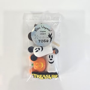 May include: A clear plastic bag containing Halloween-themed sweets. The sweets include a Mickey Mouse head, a pumpkin, and a ghost. The bag has a circular sticker with the words "Bon Voyage! From Cabin 7056" and a fairy illustration. The word "TREASURE" is printed at the bottom.