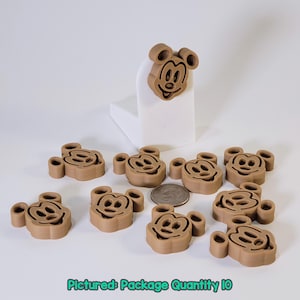 May include: A collection of ten brown Mickey Mouse-shaped embellishments. The embellishments are arranged on a white surface, with one placed on a small white block. A US quarter is included for size reference. The text "Pictured: Package Quantity 10" is at the bottom.