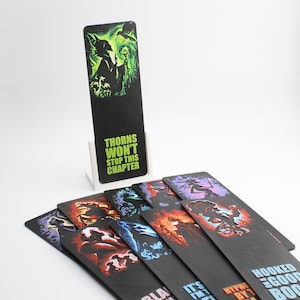 May include: A collection of black bookmarks with fantasy art. One bookmark stands in a white holder, displaying a green and black image and the text "THORNS WON'T STOP THIS CHAPTER." Other bookmarks are fanned out, each with a different design.