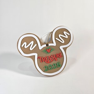 May include: A brown Mickey Mouse-shaped ornament with white icing details and the word "Treasure" in red. The year "2025" is in green. A gold string is attached for hanging. The ornament has a gingerbread cookie design.