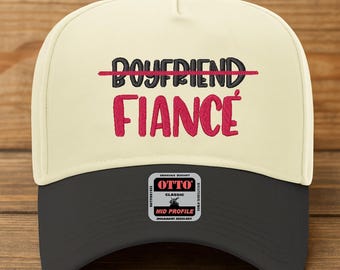 May include: A cream-colored baseball cap with a black brim. The cap features the word "BOYFRIEND" crossed out in red, with the word "FIANCÉ" embroidered in pink. A small "OTTO" patch is on the lower front.