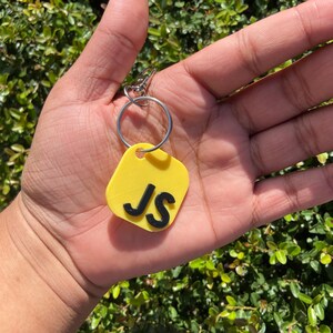 K&ouml;nnte beinhalten: Ein gelber Schl&uuml;sselanh&auml;nger mit den schwarzen Initialen "JS". Das Etikett ist rechteckig mit abgerundeten Ecken und an einem silbernen Schl&uuml;sselring und Verschluss befestigt. Der Hintergrund ist ein gr&uuml;ner, belaubter Busch.