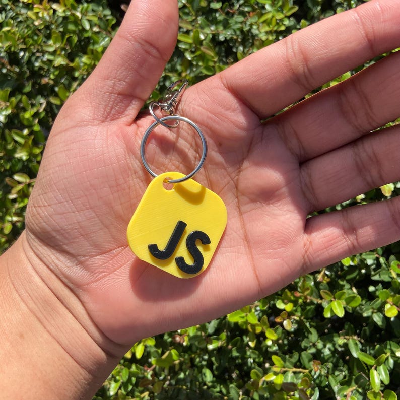K&ouml;nnte beinhalten: Ein gelber Schl&uuml;sselanh&auml;nger mit den schwarzen Initialen "JS". Das Etikett ist rechteckig mit abgerundeten Ecken und an einem silbernen Schl&uuml;sselring und Verschluss befestigt. Der Hintergrund ist ein gr&uuml;ner, belaubter Busch.